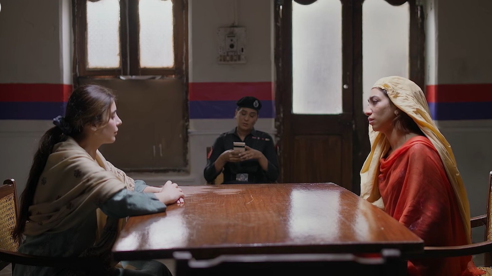 Beena and Zarmala seated across a wooden table, with a police officer behind them