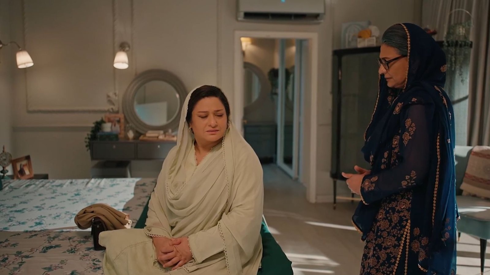 Two women, one seated on a bed in a pale dupatta and one standing in a dark dupatta